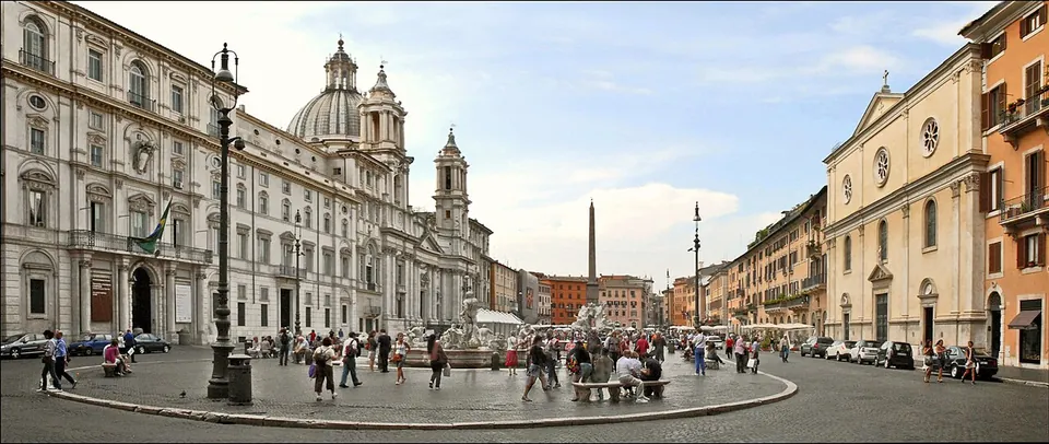 A wide panorama of Piazza Navona, a long paved square lined with palaces and a domed church, with an obelisk and fountains along its middle and people crossing it.