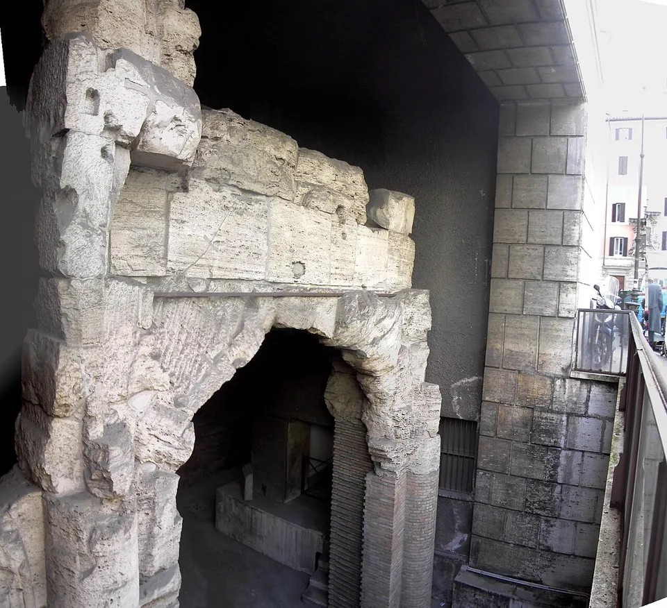 A broad arch built of large, weathered travertine blocks, its opening dark, sunk below a modern stone-clad wall with a railing and the street and buildings above it at the right.