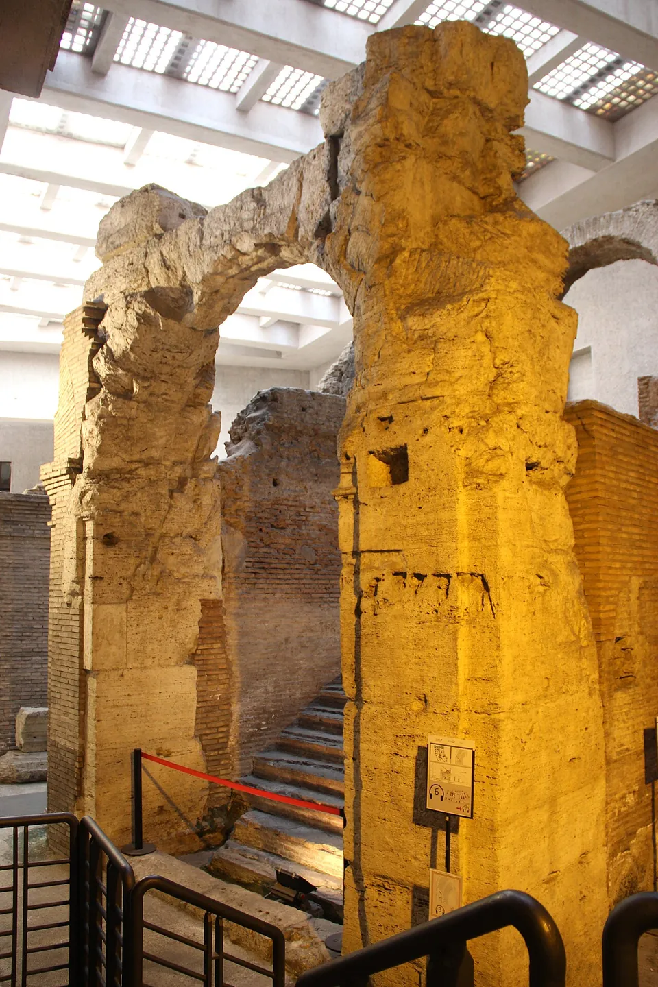 Two tall piers of travertine blocks lit warm yellow carry an arch, with a flight of steps rising between brick walls behind them and a metal railing in front.
