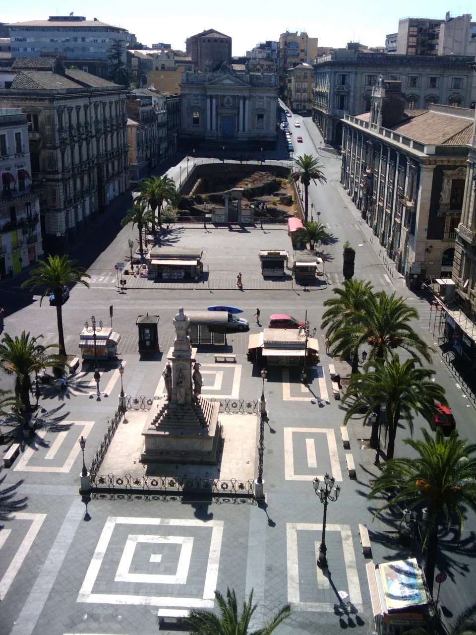 Piazza Stesicoro seen from a rooftop, a long square paved in a black and white pattern with palm trees and a monument on a plinth, and at its far end a sunken excavated area beside the road, in front of the pale façade of a church.