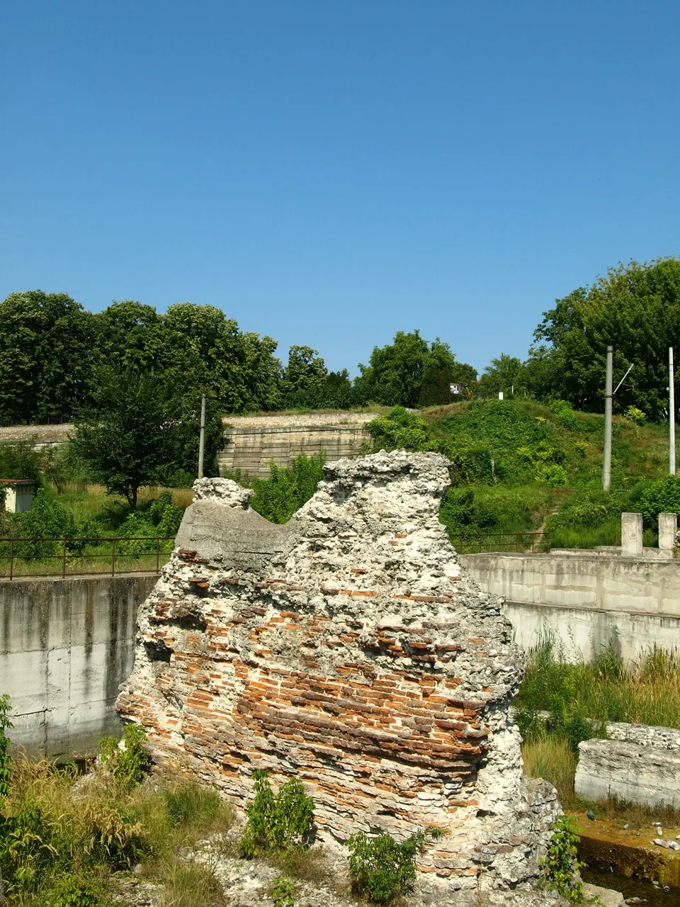 A massive weathered block of masonry, rubble and brick standing in a sunken square enclosure with concrete walls, with trees and a power line beyond.