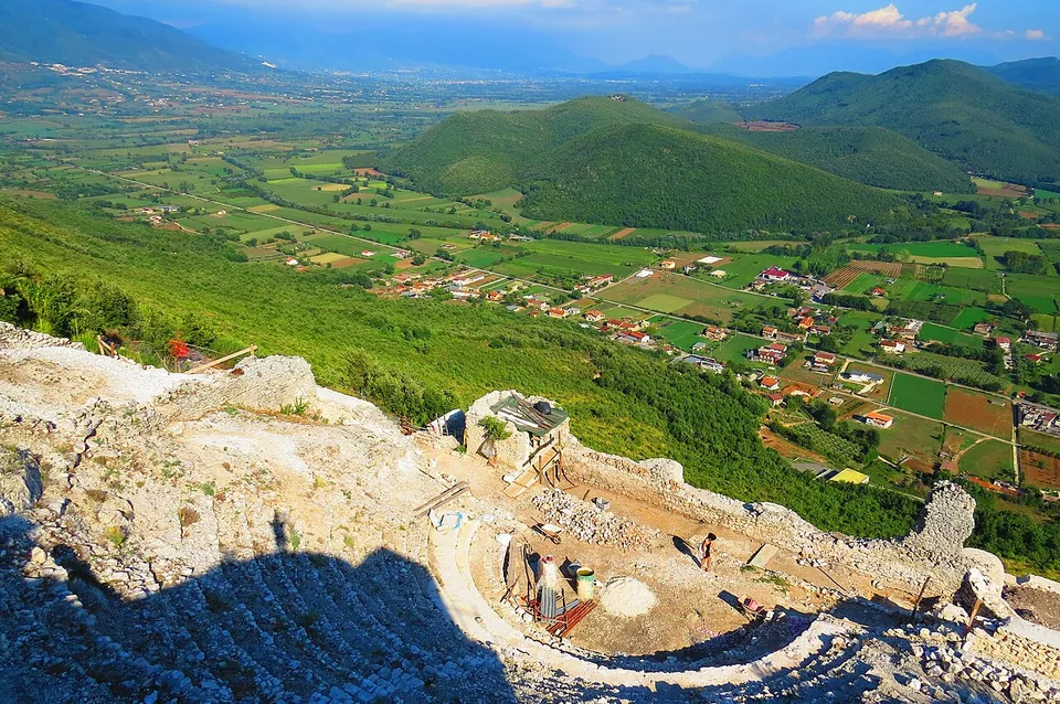 Looking down a steep wooded slope to a patchwork of fields, villages and green hills stretching to distant mountains; in the foreground the curve of the theatre's restored seating and its orchestra, with building materials stacked in it.