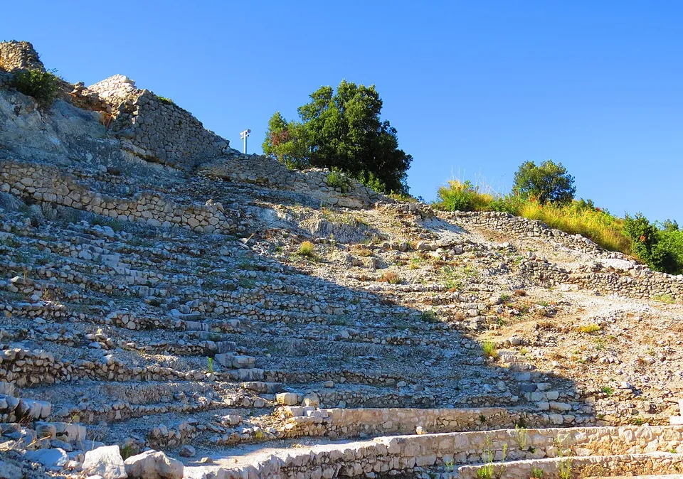 Rows of low stone steps climbing a slope, built of small rough limestone pieces set in mortar, with a broken rock outcrop and trees against a blue sky at the top.