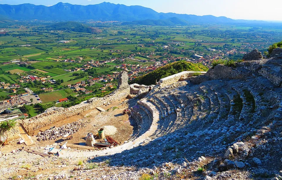 The semicircular seating of a small theatre in pale limestone rubble, rows of steps curving round a flat orchestra, with the plain, a town and a mountain range far below and beyond.