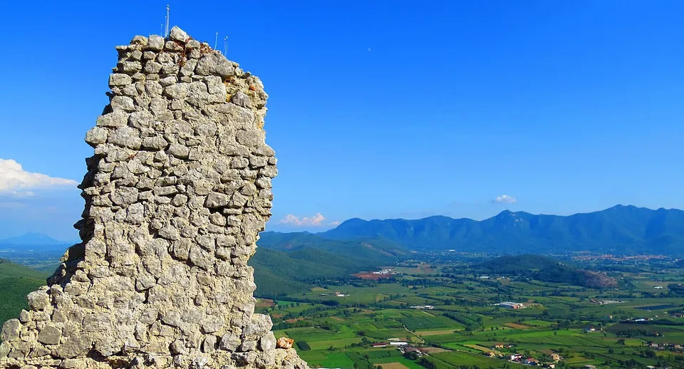 A tall, narrow fragment of wall built of irregular limestone pieces and mortar, standing alone against the sky with the valley and mountains behind it.