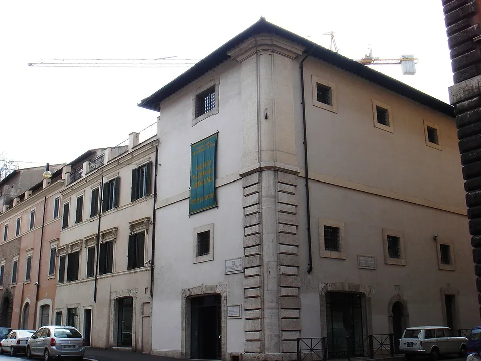 A plain three-storey palazzo on a street corner, with stone quoins at the corner, a green banner on its façade, barred ground-floor windows and cars parked along the kerb, and a building crane against the sky behind.