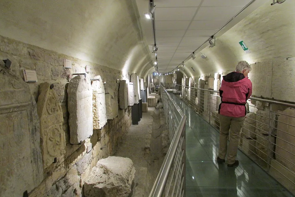 A long, low vaulted corridor lit from above, a walkway with a glass floor and steel rails running down it, and carved gravestones and stone fragments set along the left-hand wall, with a visitor reading a label.