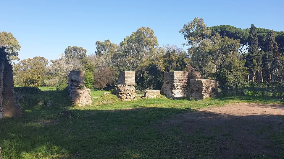 Four squat piers of brick and rubble standing in rough grass in a sunny clearing, with a dirt track and a belt of trees behind them.