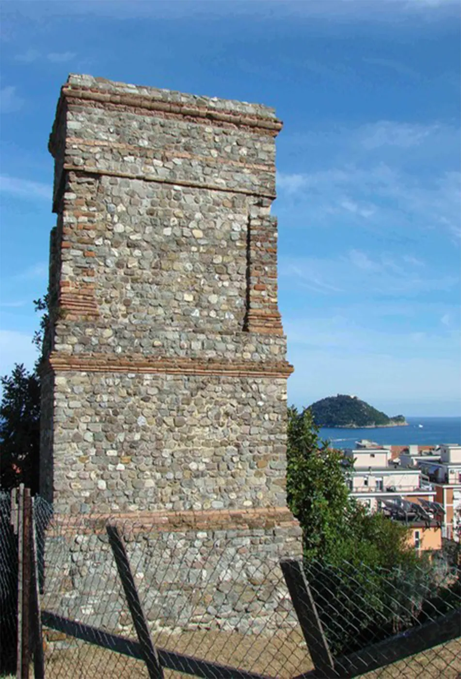 A tall rectangular tower of rubble stone with bands of red brick, behind a wire fence, with houses below and a small wooded island in the sea in the distance.