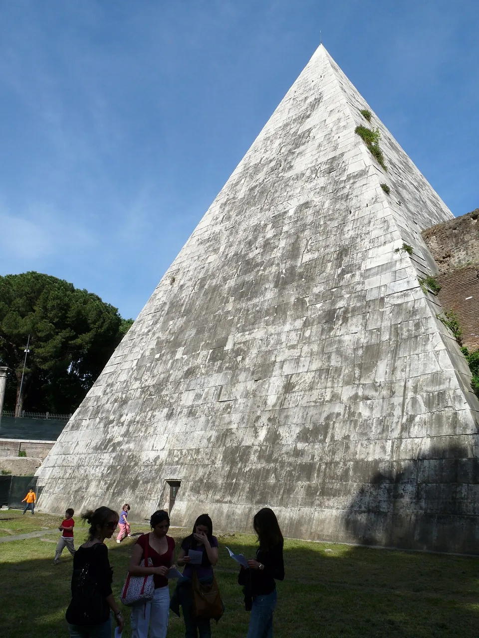 The weathered grey-white face of the pyramid rising above a lawn where a few visitors stand, with a small dark opening low in the masonry and a brick wall at the right edge.