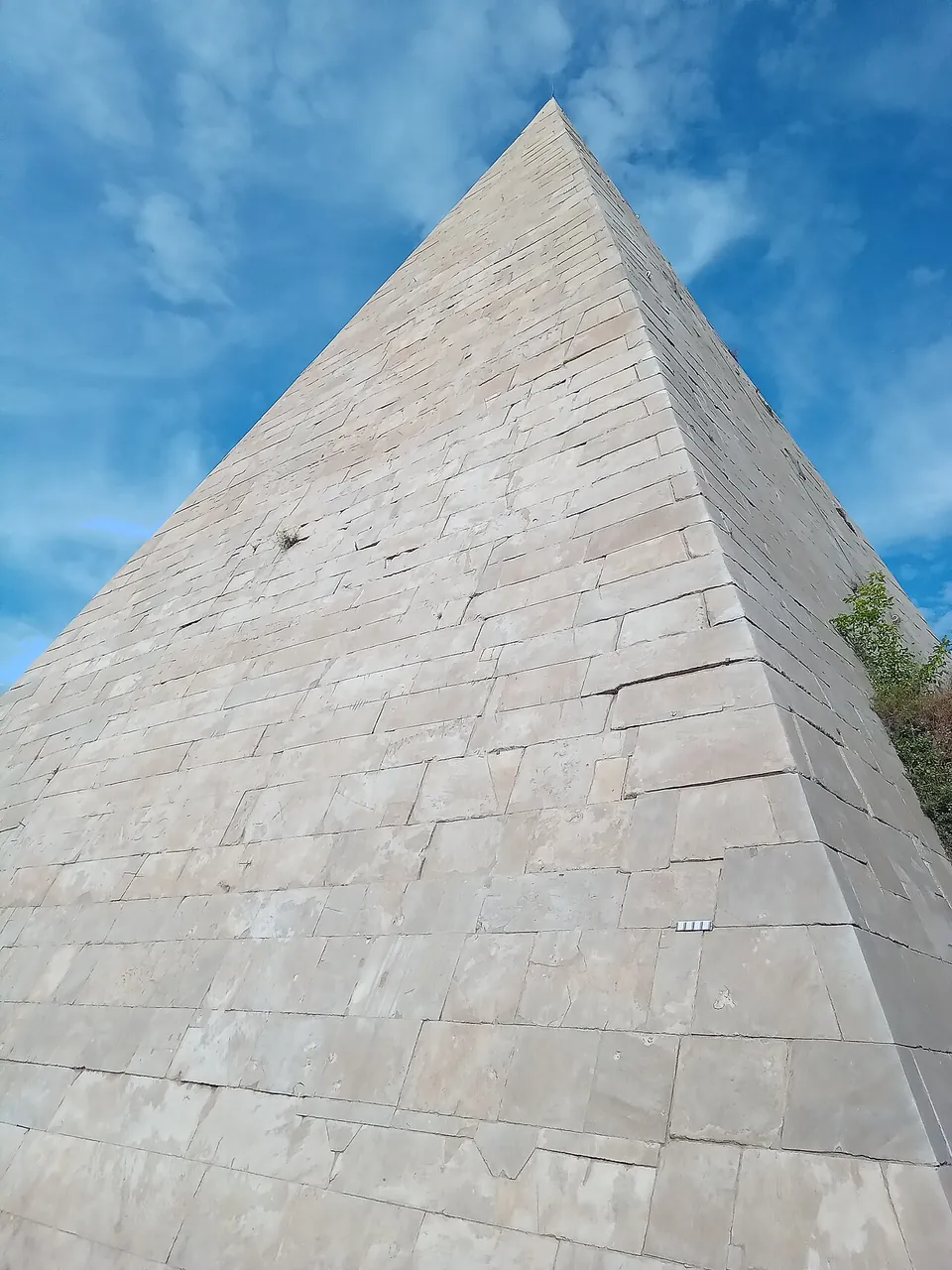 Looking straight up one edge of the pyramid, a surface of rectangular marble blocks laid in courses and narrowing towards the point against a cloudy blue sky.