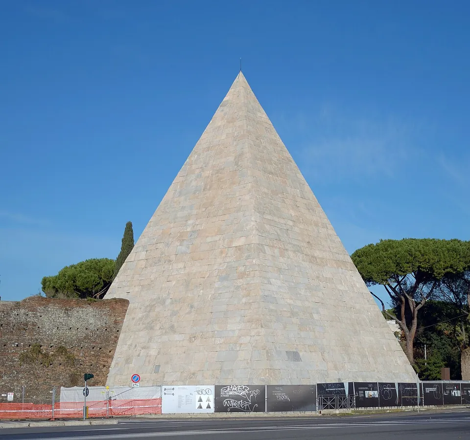 The pyramid's clean pale marble face under a deep blue sky, rising from a line of panels and fencing at street level, with the rough stone of the Aurelian Wall running off to the left and umbrella pines behind.