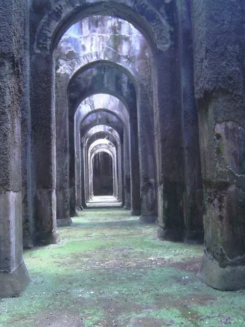A long aisle inside the cistern, a row of tall arches on massive piers receding into the distance, with damp stained walls and green moss covering the floor.