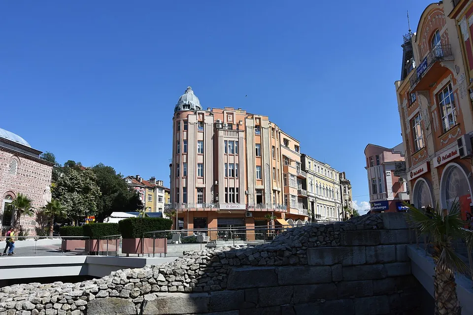 A city square under a blue sky, edged by tall early-20th-century buildings and the domed corner of a mosque, with rows of large stone seating blocks rising out of a sunken area in the foreground.