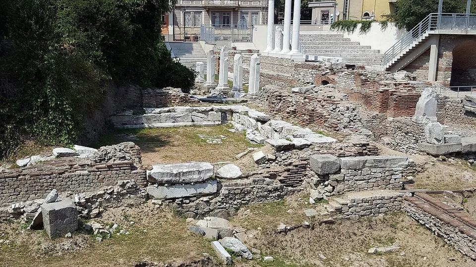 Low walls of brick and stone and scattered marble blocks in an excavated area, with re-erected columns, steps and a modern walkway in the background.
