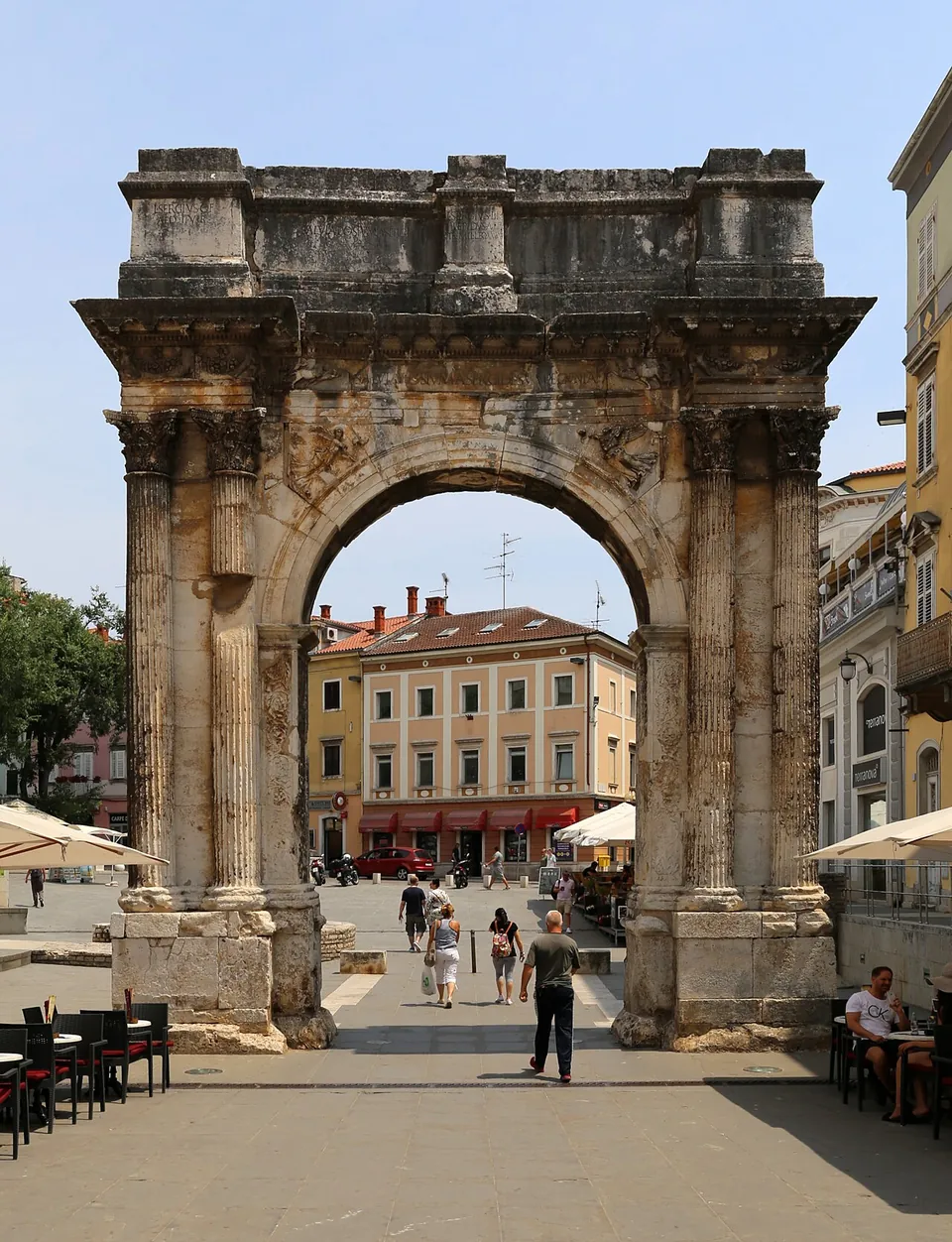 A Roman arch of pale, weathered limestone seen head on, with paired fluted columns on each side, a carved frieze and an inscription below a heavy attic, and a sunny square visible through the opening.