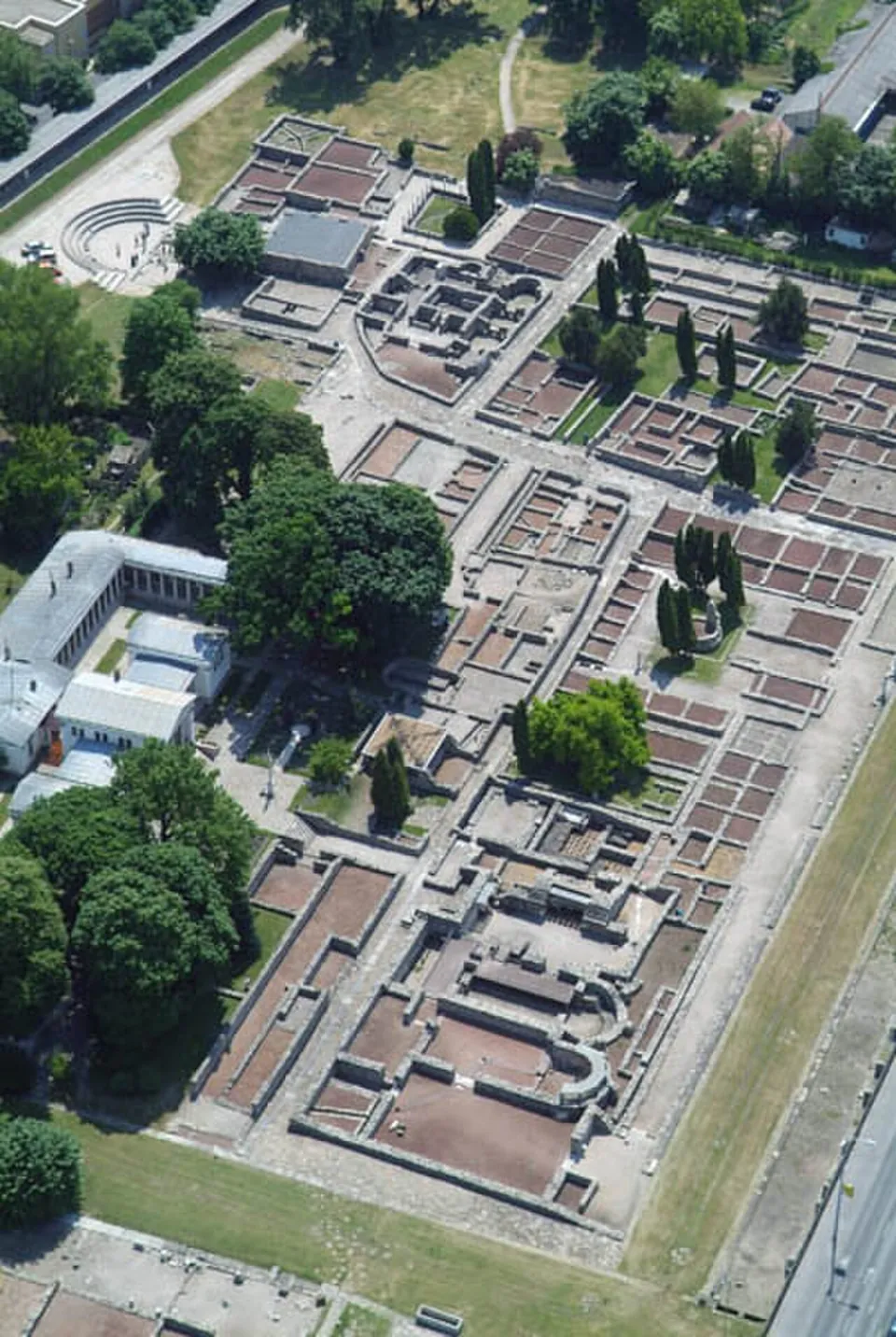 An aerial view of the excavated civil town, a grid of low stone walls and paved streets laid out across lawns, with rows of dark cypresses, low white buildings on the left and a road along the right edge.