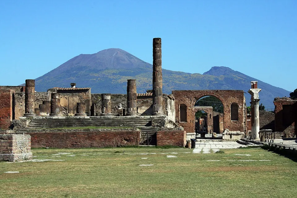 A grassy open square ending in a raised platform of brick and stone with broad steps and a few broken columns, a tall brick arch to the right, and the double summit of Vesuvius against a clear blue sky.