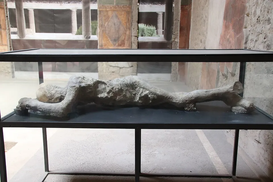 A grey plaster cast of a human body lying on its side in a glass case, in a room with painted walls, a colonnaded garden visible beyond.