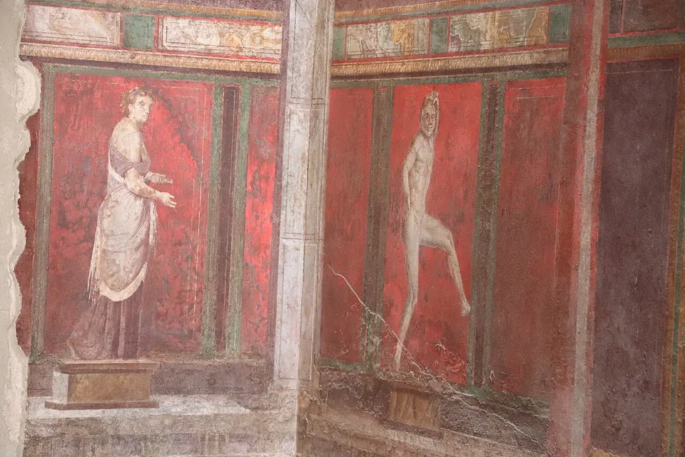 Two painted figures on a deep red wall, divided by a painted pilaster; on the left a woman in a long pale robe stands with one hand held out, on the right a naked figure steps forward.