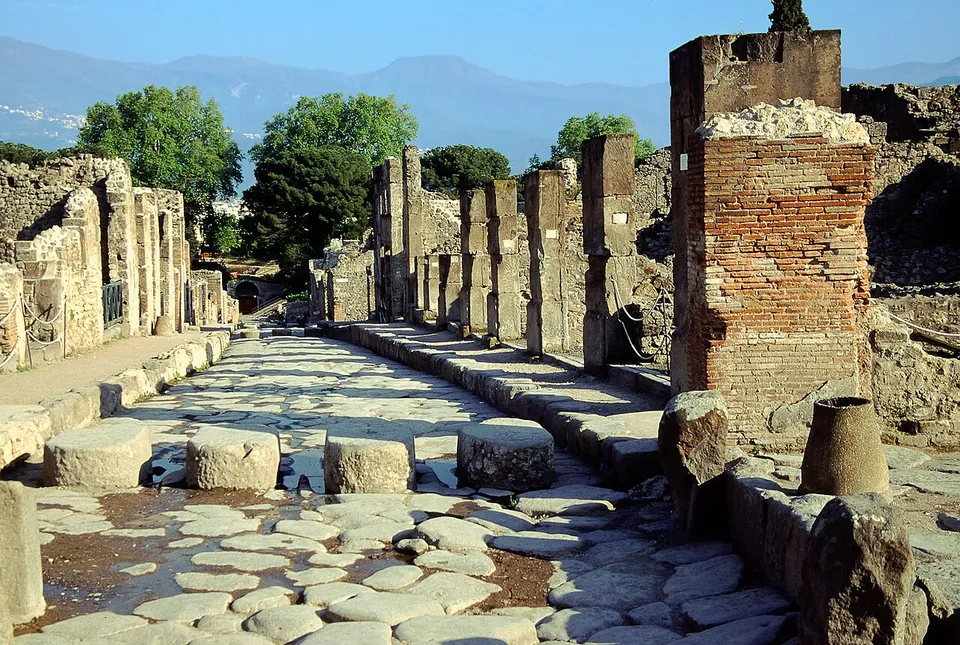A straight street paved with large polygonal stones between raised kerbs, with a row of big blocks set across the roadway and ruined walls of brick and stone on both sides, trees and hills beyond.