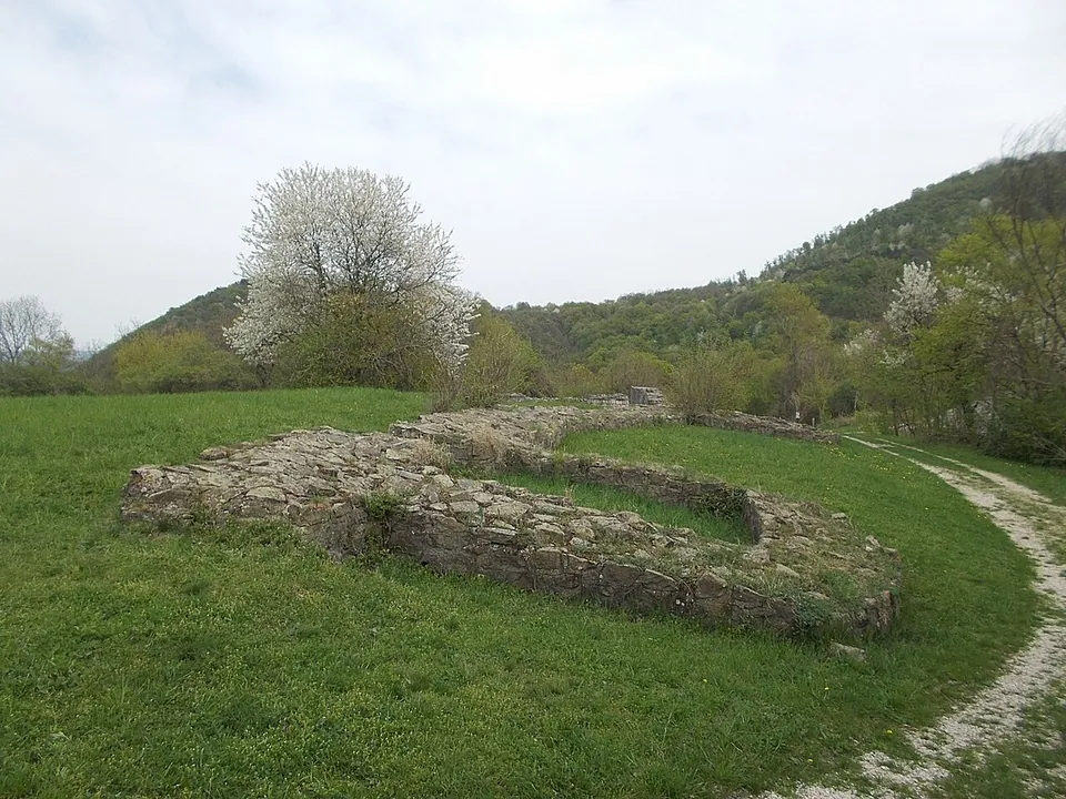 The low U-shaped foundation of a tower projecting from a straight wall line, built of rough stone and set in bright green grass, with a gravel path on the right and wooded hills beyond.
