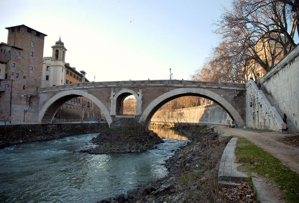 A stone and brick bridge with two wide arches and a small arched opening in the central pier, crossing a fast-flowing river to an island with a tall brick tower at the left.