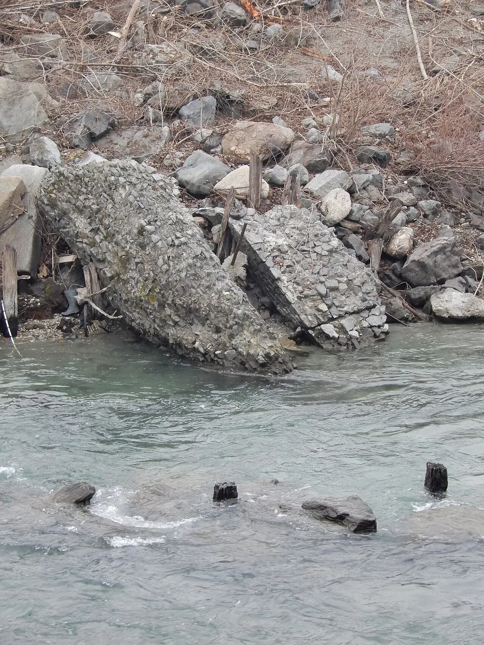 Two large tilted blocks of grey conglomerate lying among river stones at the water's edge, with a few dark stumps breaking the surface of the river in front of them.