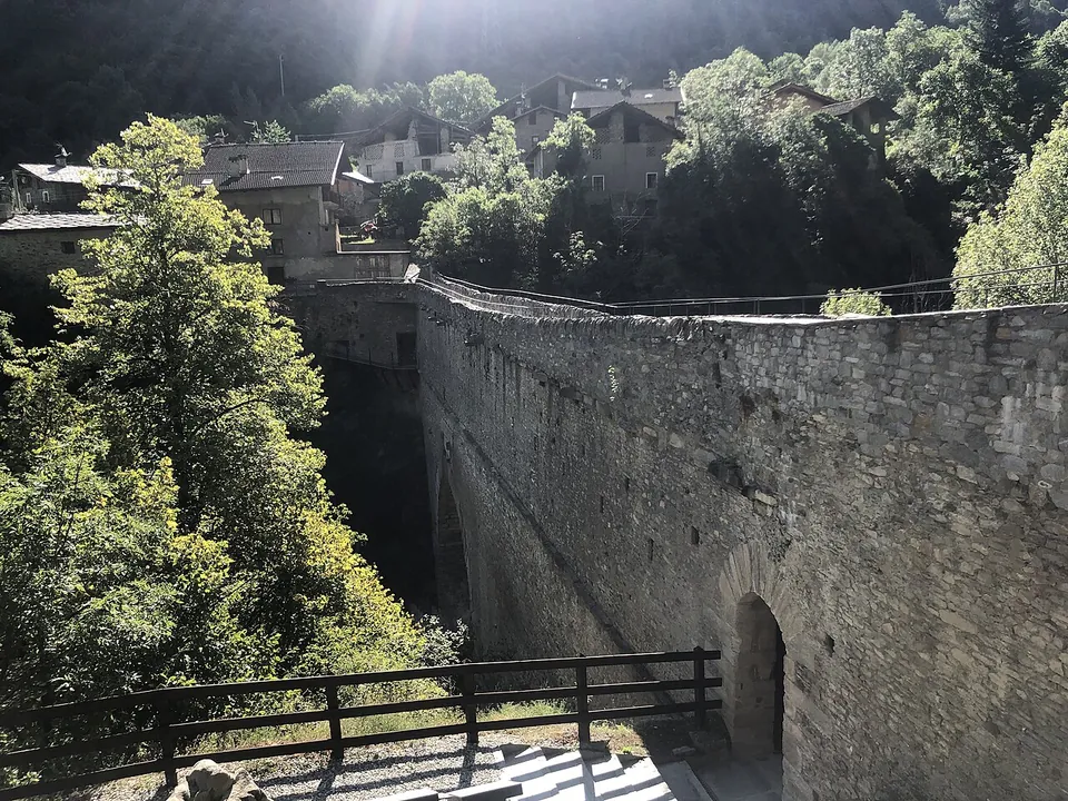 The long stone aqueduct bridge seen end-on from a terrace with a wooden railing, its arch dark over a deep wooded gorge, with the stone houses of the hamlet clustered at the far end.
