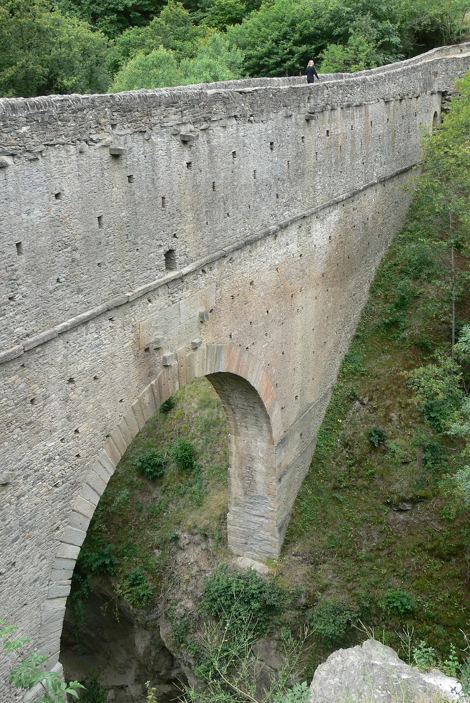 A tall, narrow wall of grey dressed stone with a single round arch at its foot, rising straight out of a wooded gorge; rows of small square openings run along its face and a person stands on the top for scale.