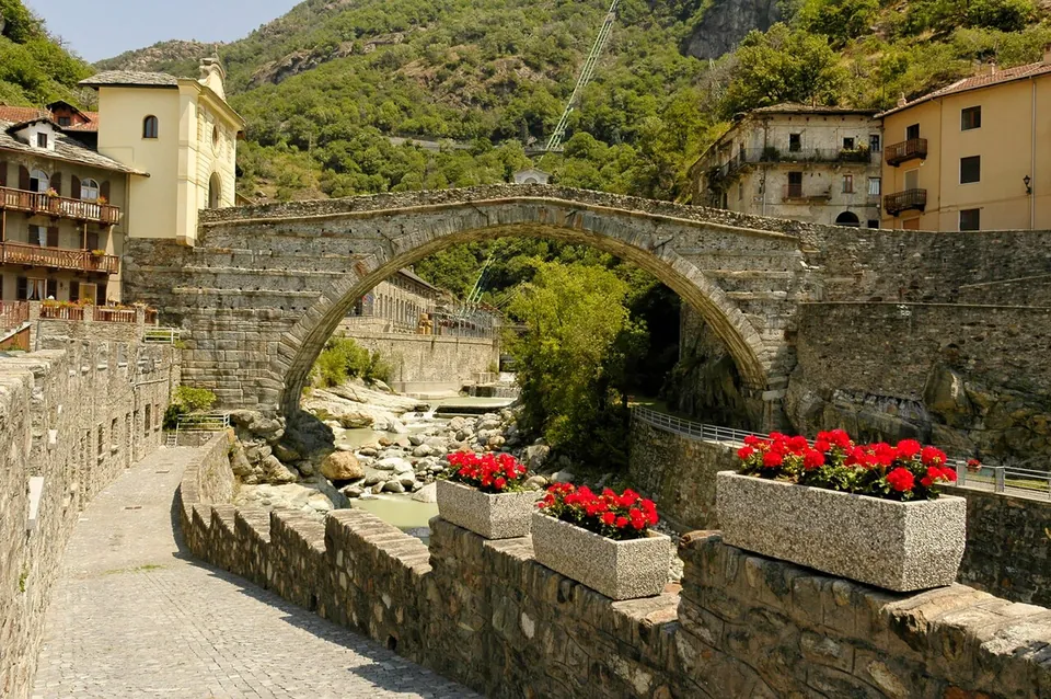 A single wide arch of grey rubble stone over a rocky streambed, between the houses of the old town, seen from a cobbled lane with planters of red flowers on the parapet in front and wooded mountainside behind.