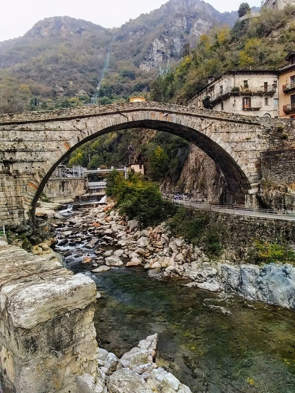 The arch seen from the rocky bed of the stream, with clear water and boulders below it, houses on the far bank and a steep rock mountain rising behind.