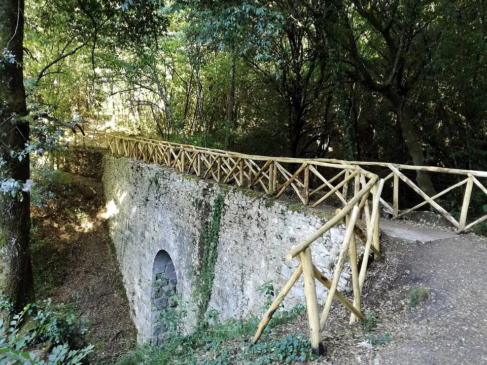A small stone bridge with a low round-arched opening, overgrown with ivy, carrying a gravel footpath with a rustic wooden railing through woodland.