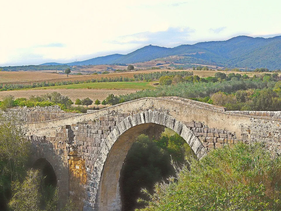 The bridge seen side-on from a height, its main arch of squared blocks and a smaller arch at the left end, with a low parapet along the deck and fields, olive groves and a mountain ridge behind.