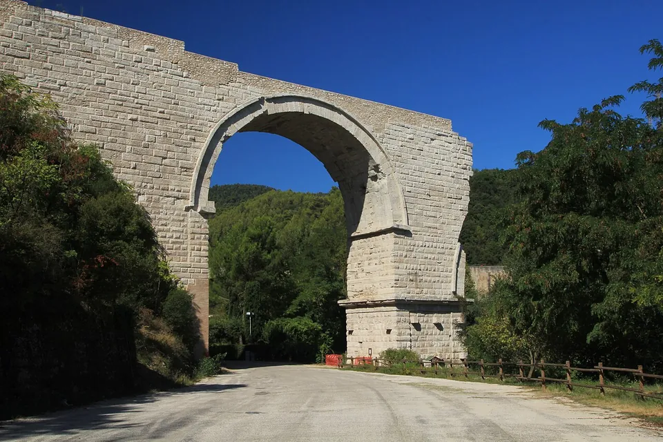 A single tall round arch of pale squared stone blocks, ending in a broken edge on the right, spanning a gravel road with a wooden fence along one side and wooded hills behind.