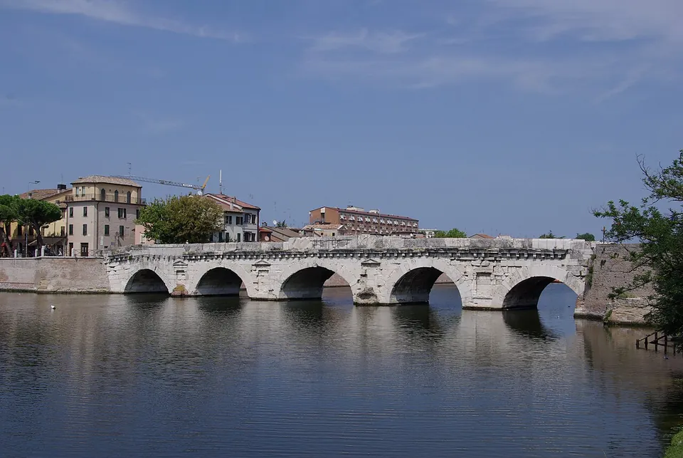 A long, low bridge of pale stone on five round arches, seen side-on across still water, with a corbelled cornice running under the parapet and houses and trees on the far bank.