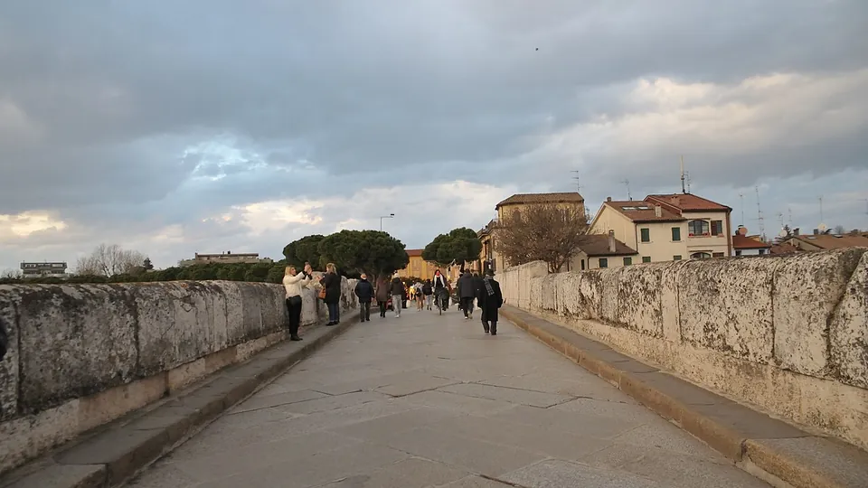 The paved roadway across the bridge between two parapets of large rounded stone blocks, with people walking across towards low houses and trees at the far end.