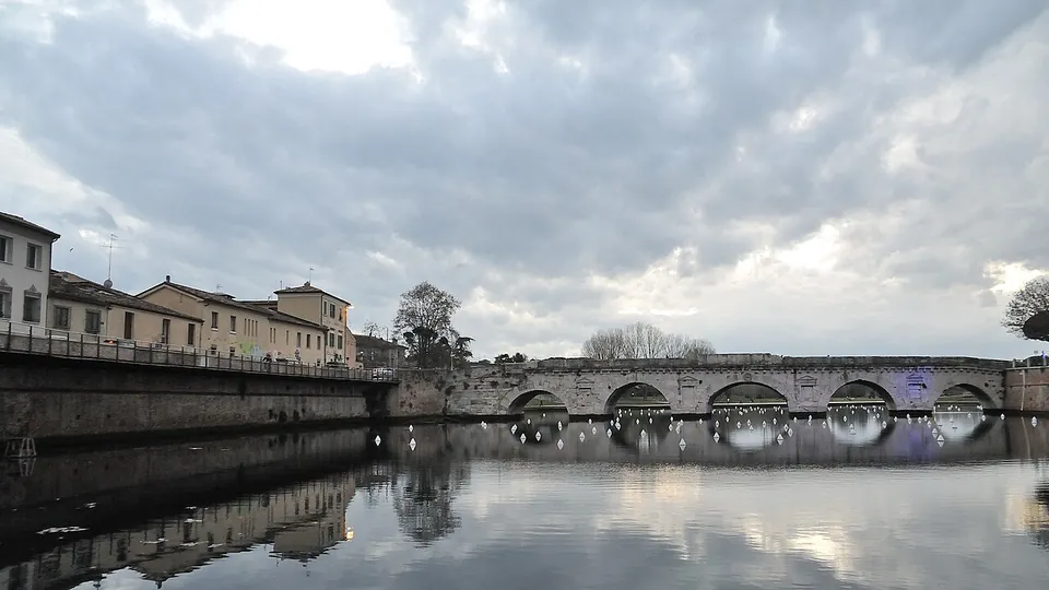 A broad sheet of still water under a cloudy sky, with brick quay walls and a row of houses on the left and the low stone bridge closing the far end of the basin, its arches reflected in the water.