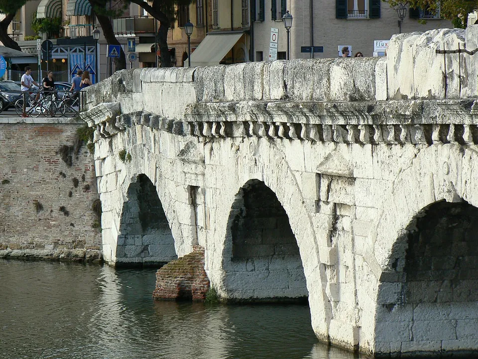 Close view of three stone arches rising from the water, built of weathered white blocks, a shallow niche with a small pediment carved on the pier between two arches, and a row of projecting brackets carrying the cornice under the parapet.
