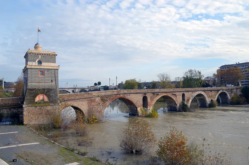 A stone bridge of round arches over a muddy, fast-flowing river, its piers pierced by smaller arched openings, with a square stone tower topped by a small cupola and a flag at the near end.