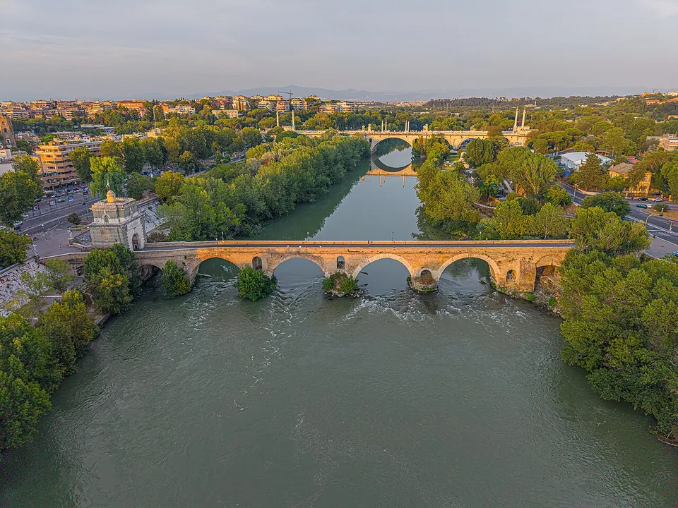 An aerial view along a broad green river in evening light, crossed by a long stone bridge of several arches with a square white tower at its left end, wooded banks and apartment blocks on either side, and a second bridge farther along the river.