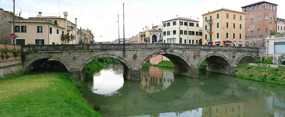 A long, low stone bridge of five flattened arches crossing a green river, with grassy banks in front and a row of pastel-coloured town houses behind.