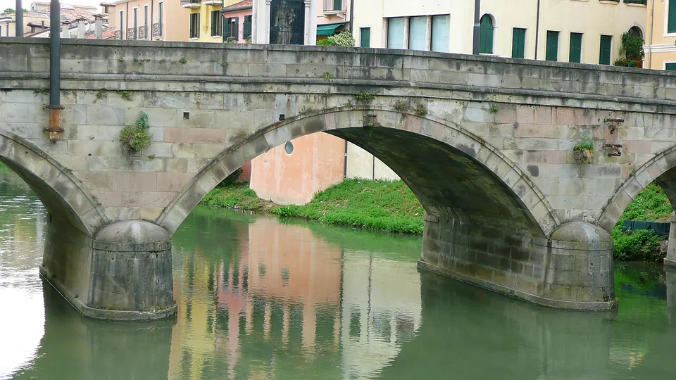 Close view of two of the bridge's flattened stone arches resting on a pier with a rounded cutwater, reflected in still green water.