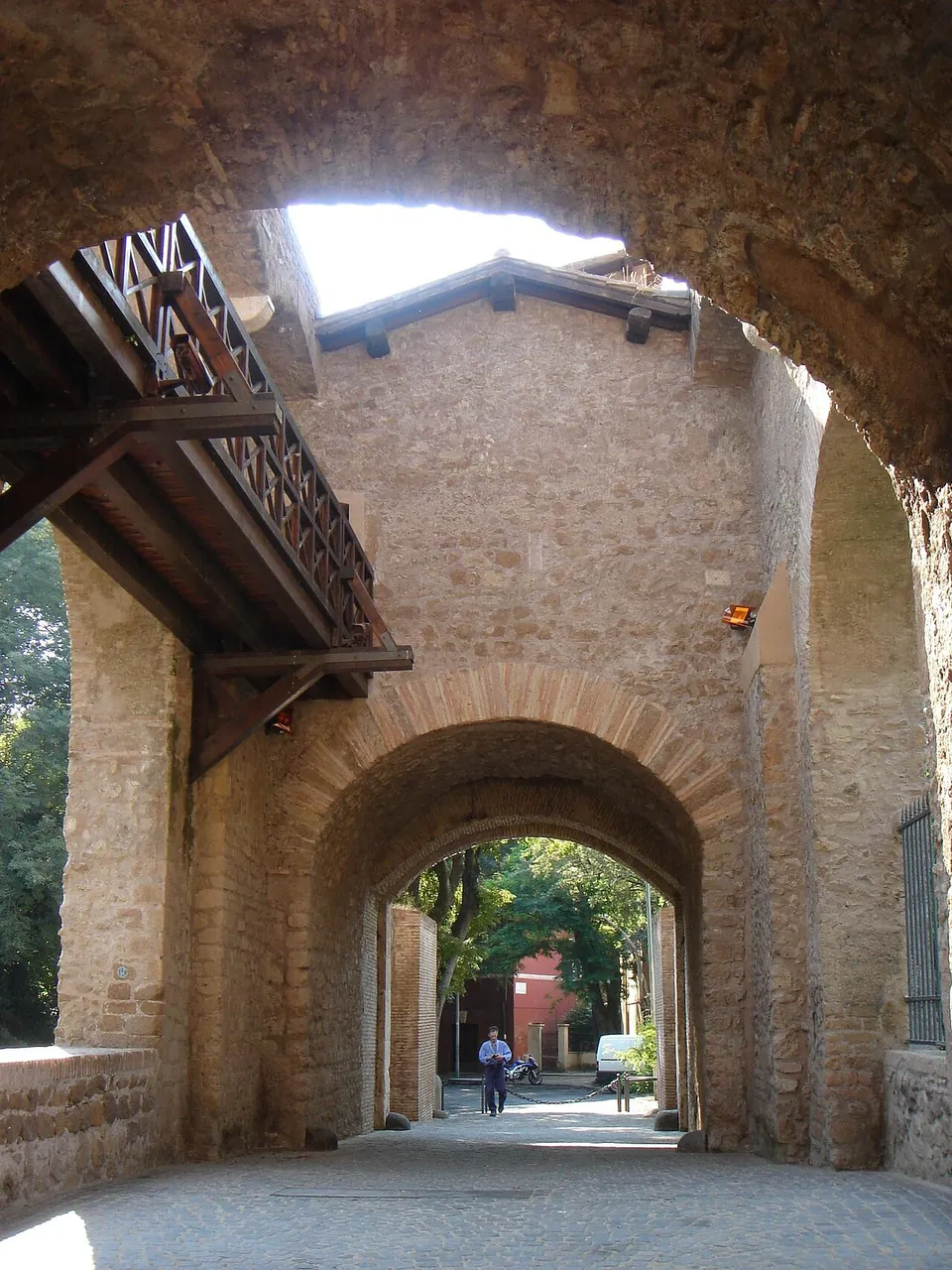 Inside the bridge, a paved passage runs between masonry walls under a wooden balcony to a second round arch, with trees and a building beyond.