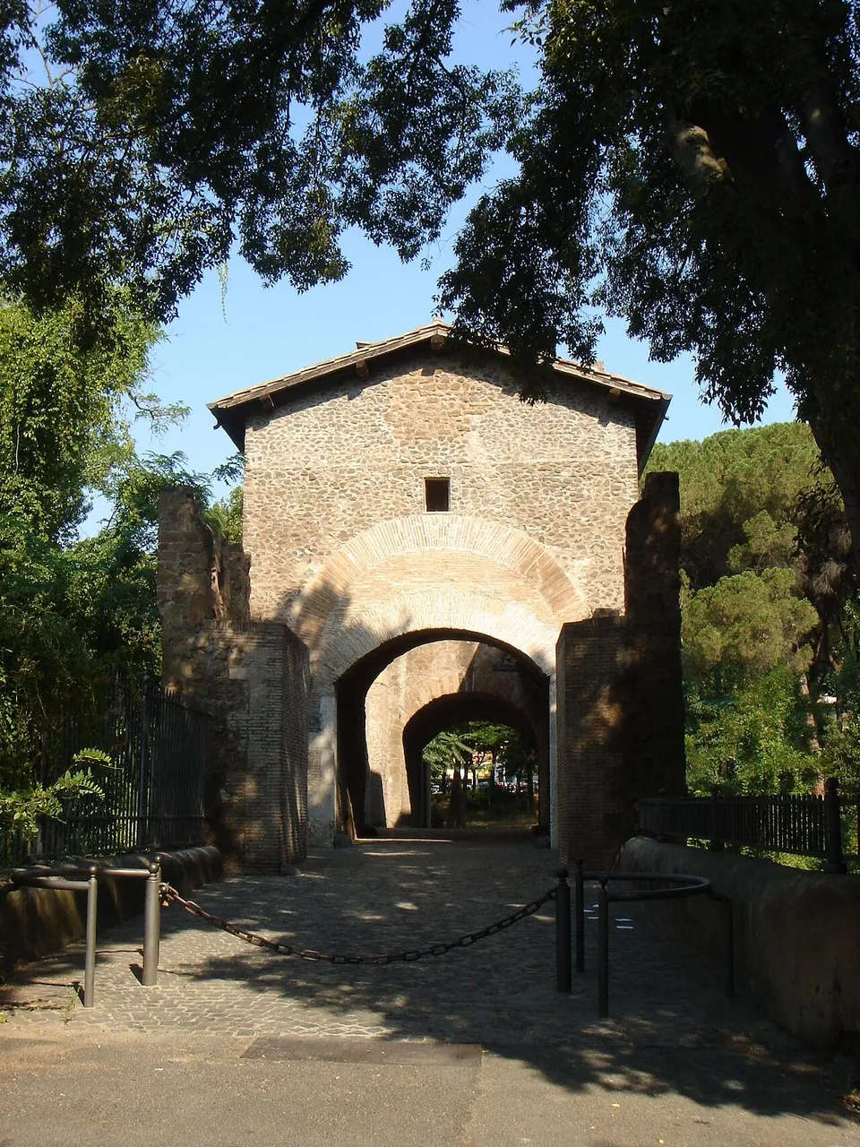 A cobbled path leading under trees to the gateway of the bridge, a tall stone block with a pitched roof and a round-headed arch, through which the far end of the passage is visible.