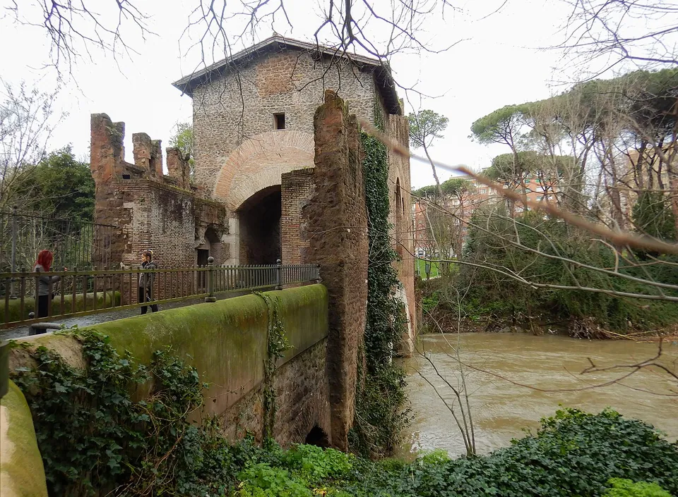 The side of the bridge above a swollen brown river. Two people stand at a railing on the walkway, and behind them rise crenellated brick walls and a square tower with a large arched opening, with ivy climbing the stonework below.