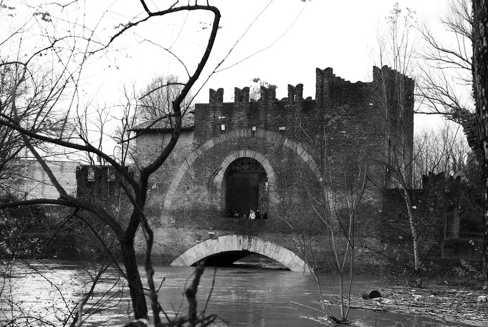 A black-and-white view from the riverbank through bare winter branches. A single wide arch spans the water low down, and on top of it stands a crenellated, castle-like block with an arched gateway in its face.