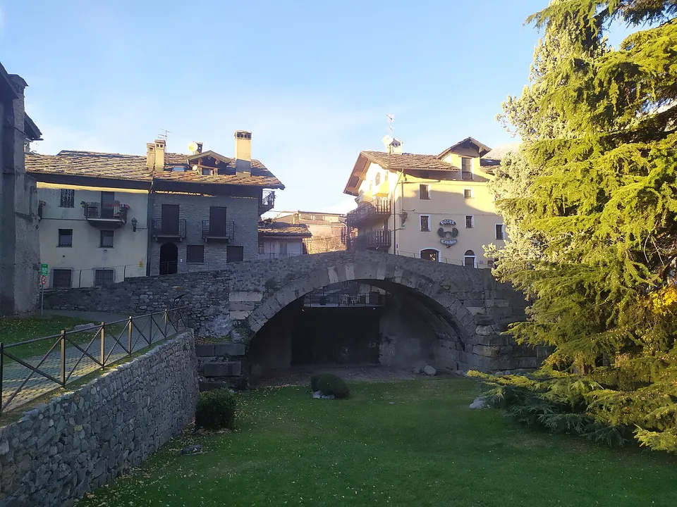 A single low stone arch spanning a dry, grassy hollow, with a railing along a path on the left, a tall conifer beside it and old houses with stone roofs behind.