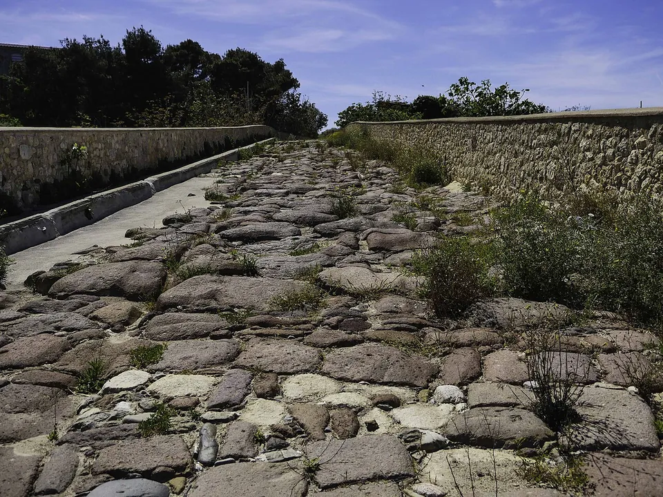 The bridge's carriageway paved with large irregular dark stones, weeds growing in the joints, running between two low parapet walls under a blue sky.