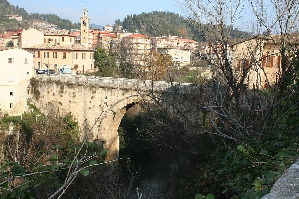 A single wide arch of pale squared stone blocks spans a deep, overgrown river gorge, carrying a road with houses and a bell tower on the far bank.